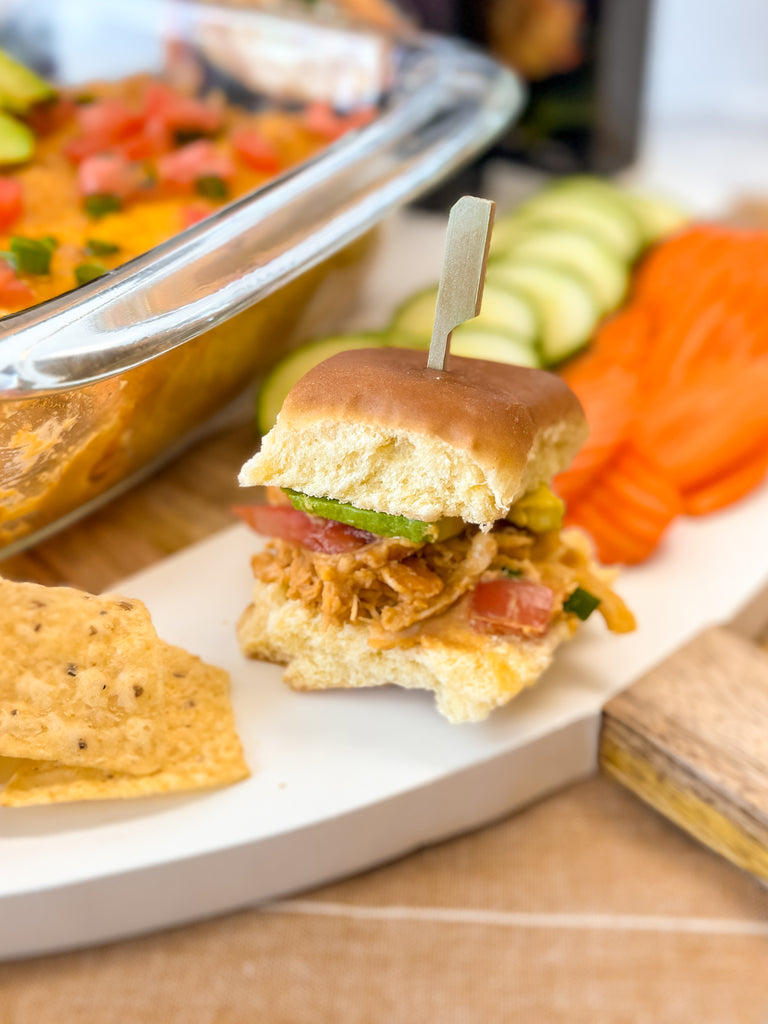 spicy buffalo chicken hot hummus dip on a slider bun with tomato and avocado sits on a board with tortilla chips and cucumber and carrot slices. The pan of hot dip can be seen in the background.