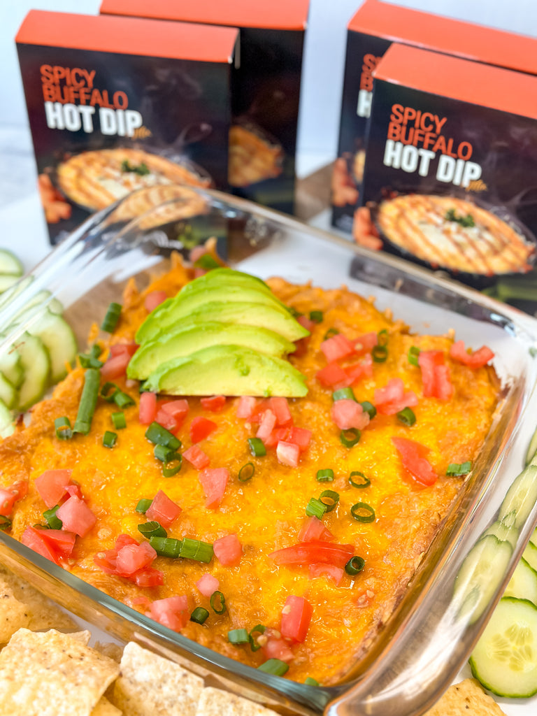 an 8x8 dish of spicy hot hummus dip on a tray with cucumber and carrot slices and tortilla chips with boxes of wind and willow spicy buffalo dip.