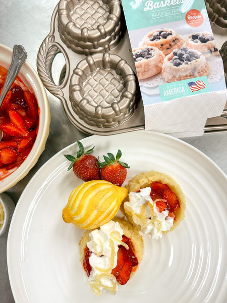 strawberry shortcakes with whipped cream and candied lemon peel on a platter with strawberries and lemon. the nordicware basket pan is in the background.