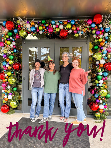 A group of four people stand in front of a Holiday decorated door at The Kitchen Store. The decor is made of green lighted garland with various sized, colorful ornaments. Text on the graphic reads "thank you."