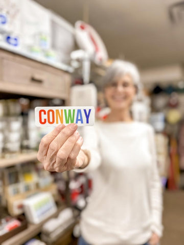 a person holds a "Conway" vinyl sticker up to the camera. Each letter in "Conway" is a different color.
