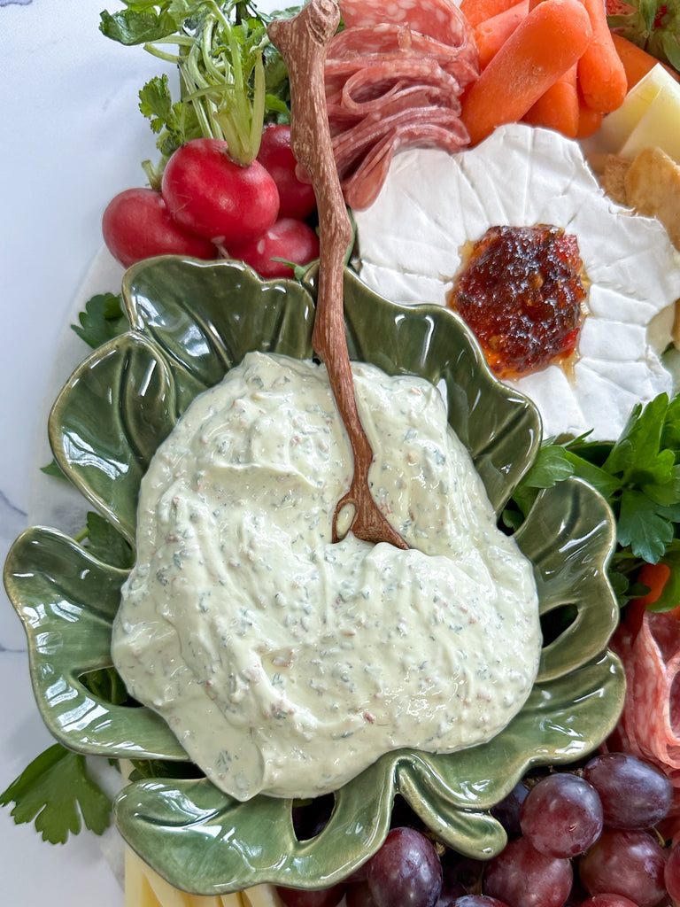 avocado dip served in a leaf shape bowl with a twig-like wooden spoon surrounded by fresh vegetables.