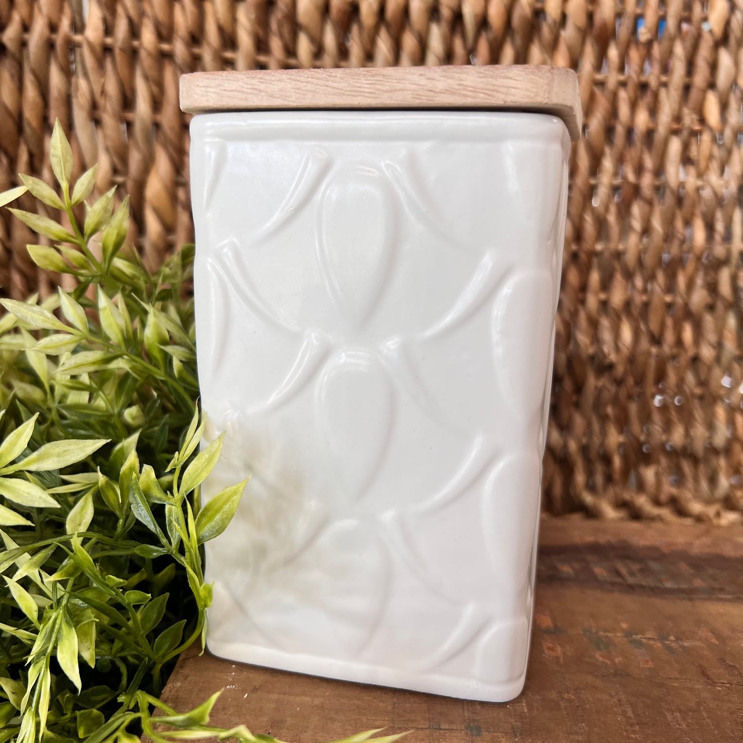 tall square candle in white ceramic canister with geometric pattern and a wooden lid set on wood table with greenery.