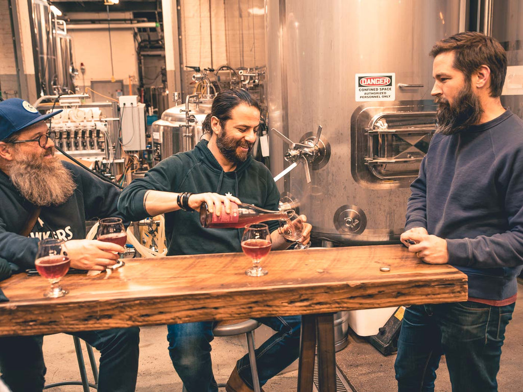 Sam from Other Half stands at the end of the table while Mazen pours an Other Half sour, seated next to Alex