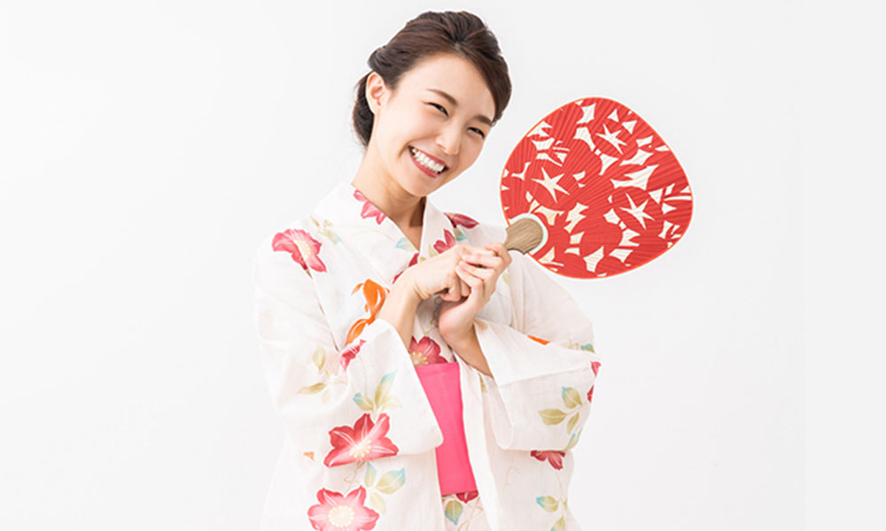 Une femme tient dans la main un éventail japonais Uchiwa de couleur rouge. Elle est habillée avec un kimono pour femme à fleur. Elle sourit et est heureuse.