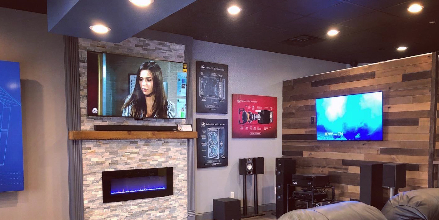 About Us – AC Audio Video Showroom with a flatscreen tv hanging on a stone wall with sound bar over a fireplace, beige leather couch and another flat panel tv on wall with free standing speakers and audio components on side wall. 