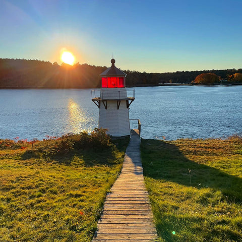 white lighthouse with red glass, sun in the background, wooden walkway leading to lighthouse