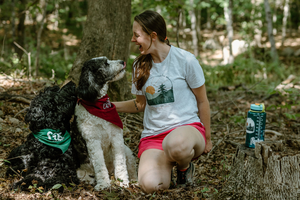 Woman sitting on ground in the forest with two dogs, with canteen water bottle sitting on stump