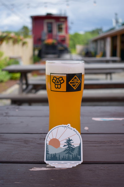 a beer with a sticker with mountains and trees that says explore more leaning on it