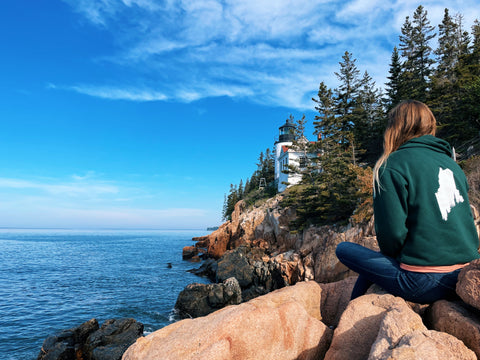 woman sitting on rocks looking at white lighthouse sitting among trees, blue sky and ocean