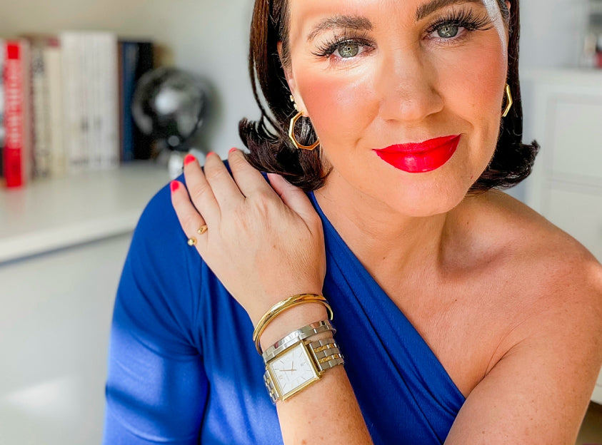 Woman in a blue dress with red lipstick, gold jewelry, and a watch, posing indoors.