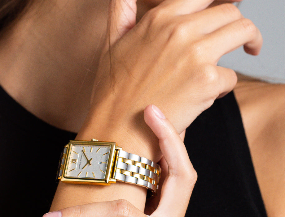 Person wearing a gold and silver rectangular wristwatch with a black strap.