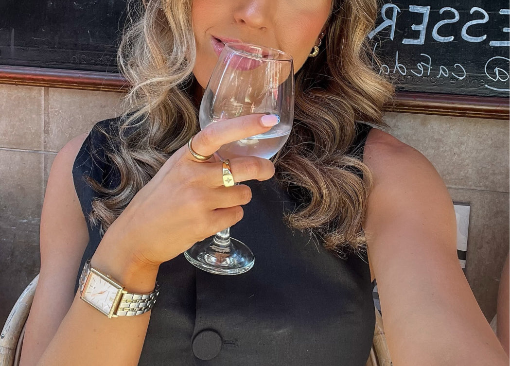 Person with curled hair holds a glass of clear liquid, wearing a watch and rings.