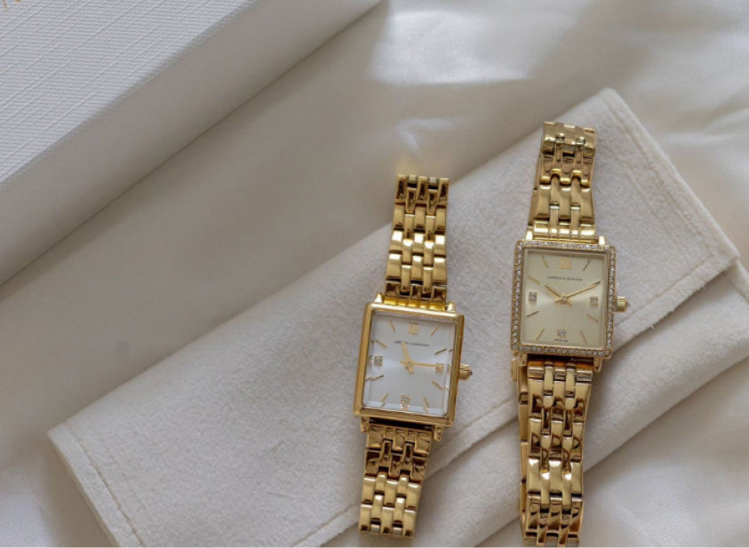 Two gold wristwatches with rectangular faces on a white cloth.