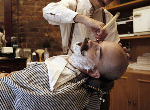 Old fashioned barber giving a straight blade shave to bald customer with handlebar mustache