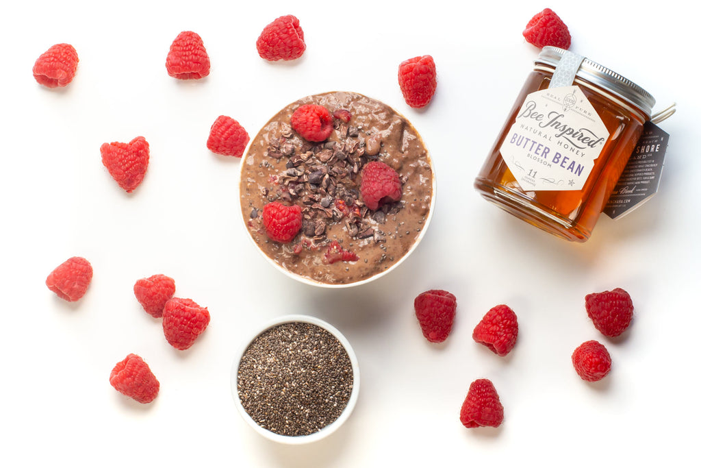 chocolate chia seed pudding on a white surface surrounded by raspberries and a jar of honey