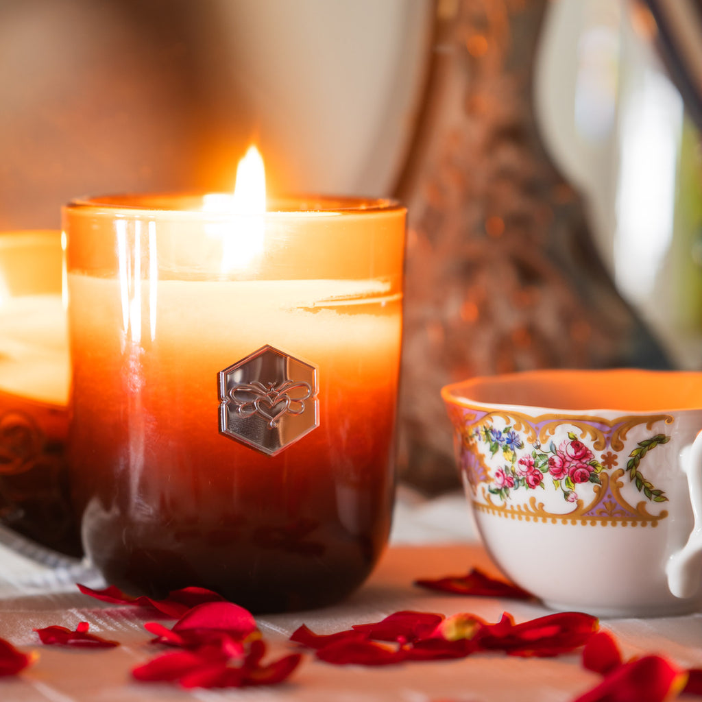 Bee Inspired Luxe Candle lit on a table in front of a mirror, next to a full tea cup, and surrounded by red rose petals