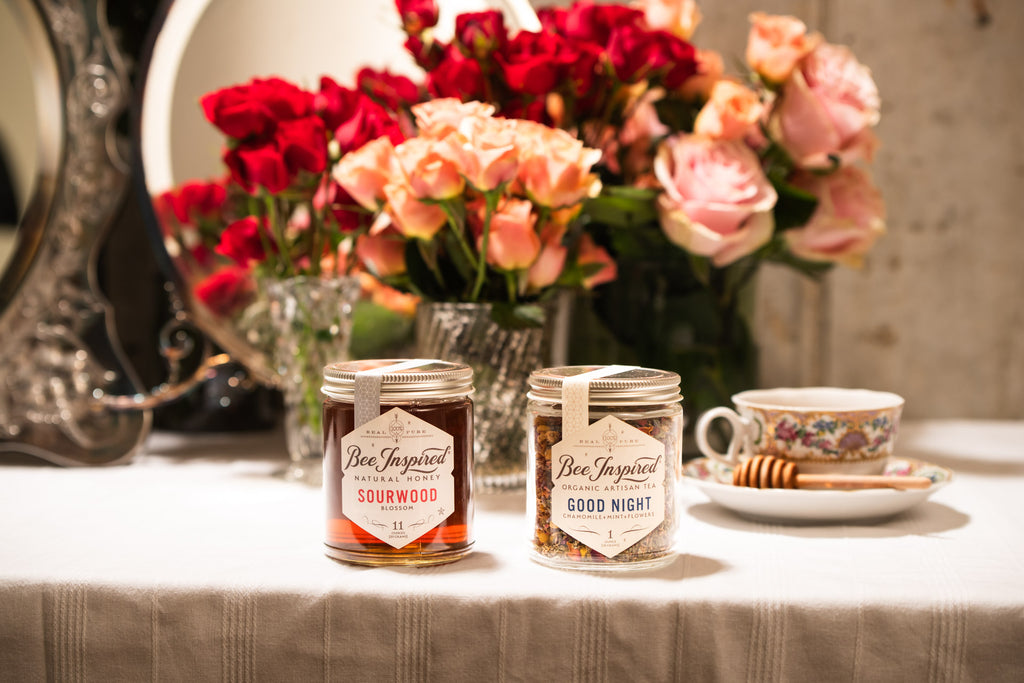 Bee Inspired Sourwood Honey and Good Night Tea set up next to each other on a table in front of a mirror and red and pink roses