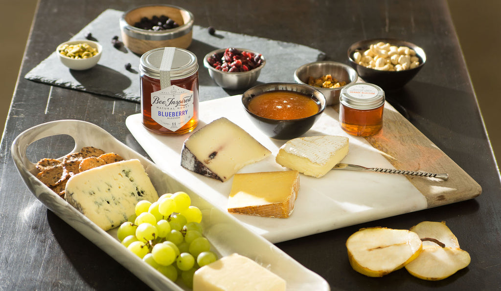 A charcuterie board spread with various cheeses, fruits, and Bee Inspired Honey.