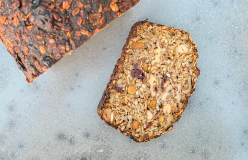 A slice of honey nut and seed bread on a light, hard surface next to the loaf.