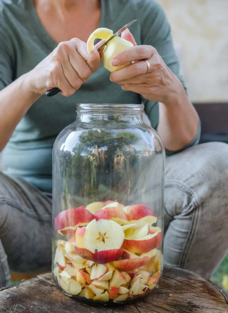 A woman sitting behind a large glass jar holding peeled apples while she is peeling an apple.