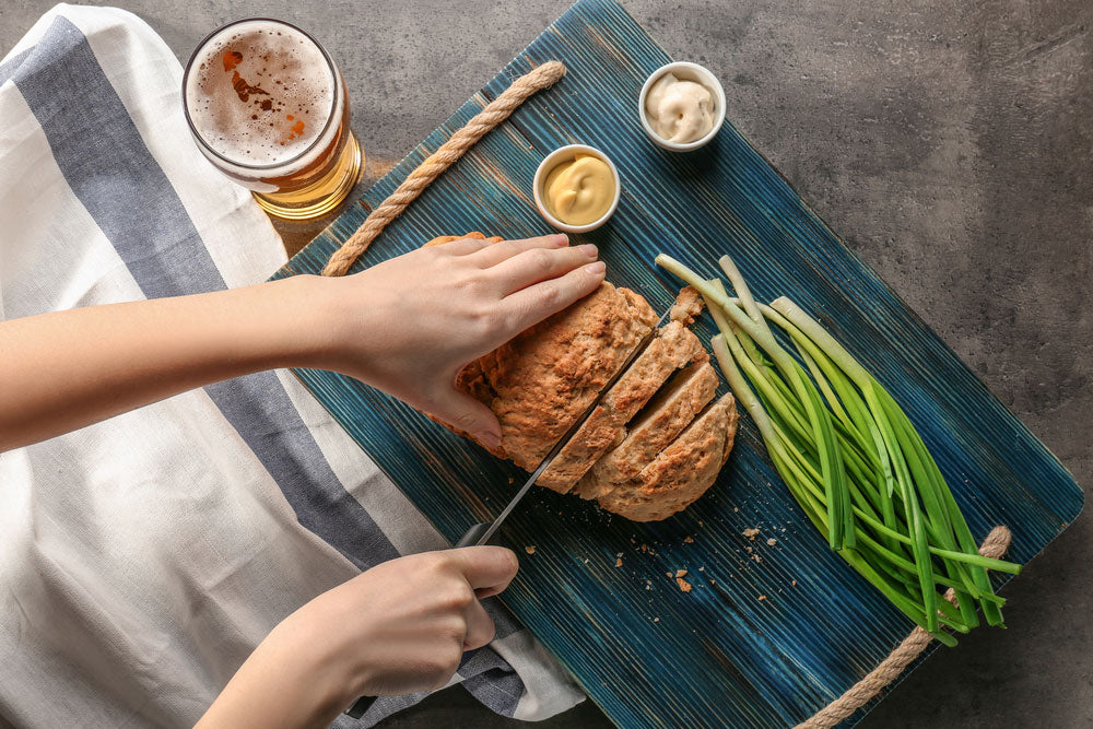 Hands slicing bread on top of a cutting board along with beer, onions, and condiments on the side