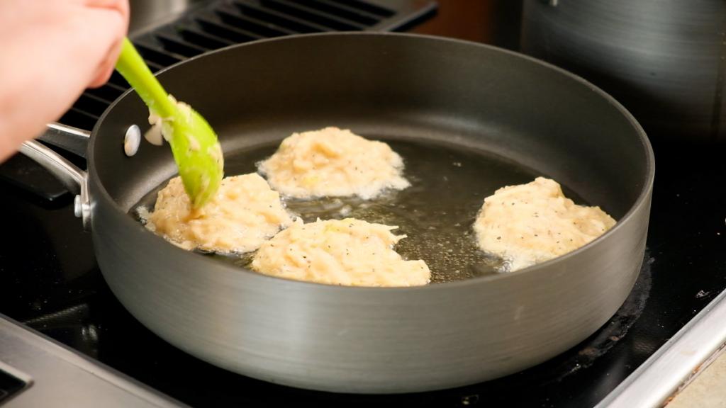 Frying latkes in a skillet, puffs of flour and oil cobwebbing out onto the  stovetop. The cook flipping the potato pancakes with a spatula, kids  eagerly peeking inside the skillet. Generative AI