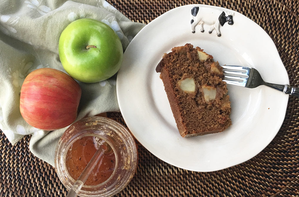 apple honey cake on cow plate with fork on wicker placemat with 2 apples a napkin and raw honey in hive container