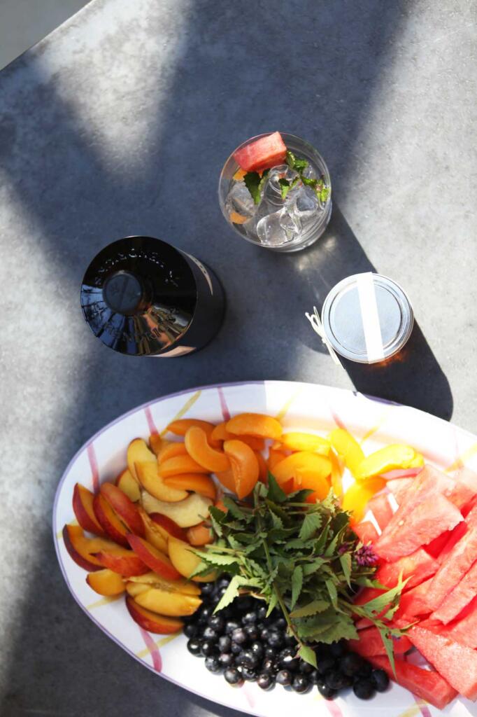 Bee Collins set with a muddled gin drink in glass, bottle of gin, jar of honey, fresh platter of fruits and mint