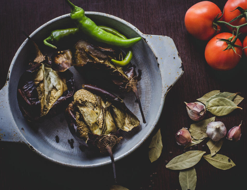 Chili roasted eggplant ingredients in pottery on dark table with bay leaves garlic cloves and tomatoes