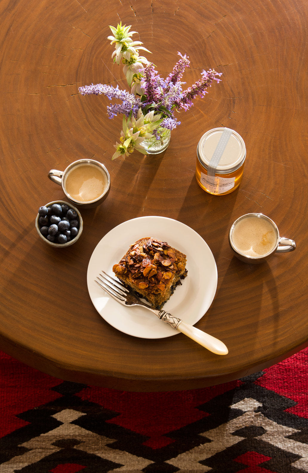 Healthy coffee cake set with jar of honey, wildflower arrangement, fresh berries in bowl, cup of espresso, with coffee cake on white plate with fork on tree stump table with navajo rug on floor
