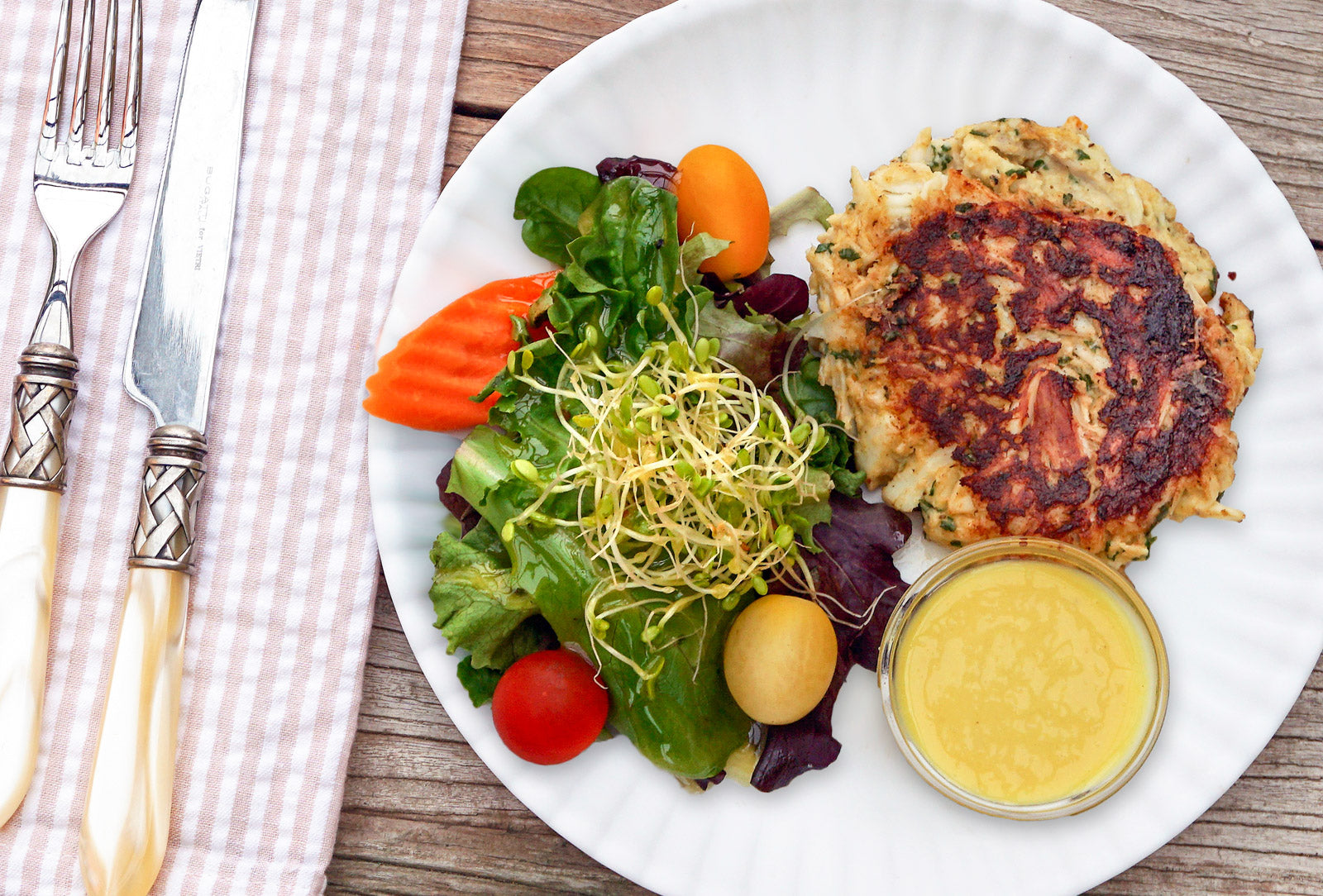 Crab cake on plate with fresh green salad and honey mustard dipping sauce on wood table with striped napkin