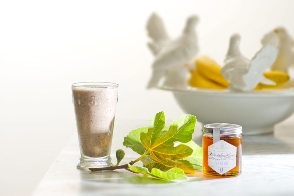 Honey Fig Smoothie in glass with fresh-picked fig leaf and fig in process on white marble counter with white bowl filled with bananas