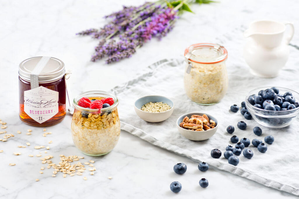 Simple overnight oats in weck jar topped with eastern shore honey and fresh berries on marble table with raw ingredients, fresh-picked blueberries, milk pitcher with napkin and fresh cut lavender.