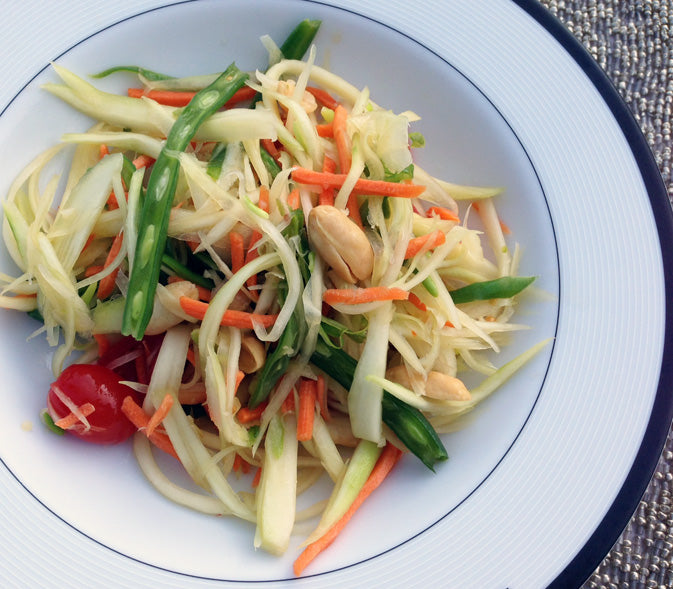 thai green papaya salad with green beans, tomatoes, peanuts, with a tangy pungent chili lime dressing in white bowl on placemat