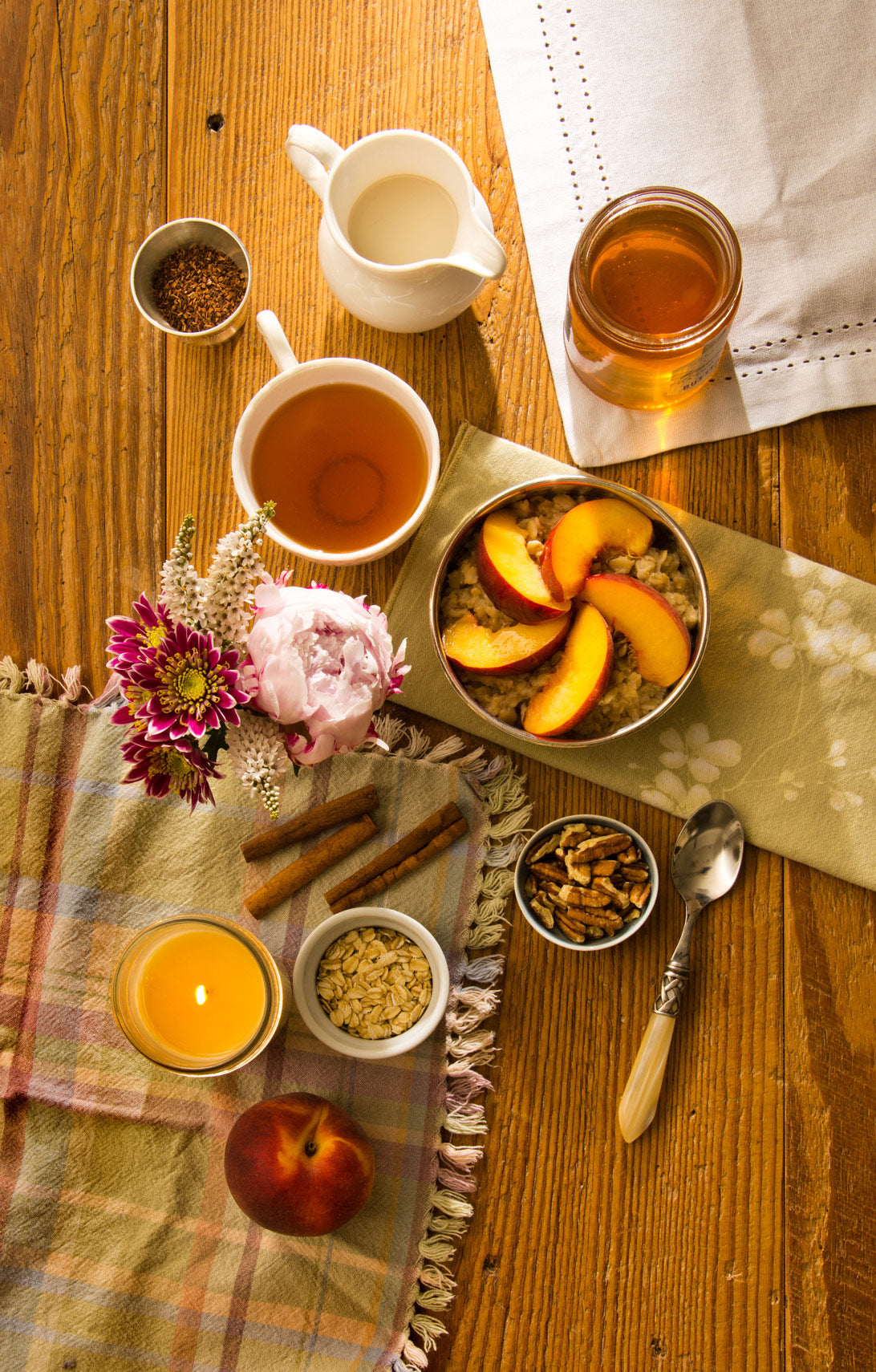 Warm Peach Oatmeal served with fresh peach slices and eastern shore honey with milk pitcher and side of nuts. Candle burning. Napkins and placemats and raw ingredients on the table with hot tea and bouquet of flowers