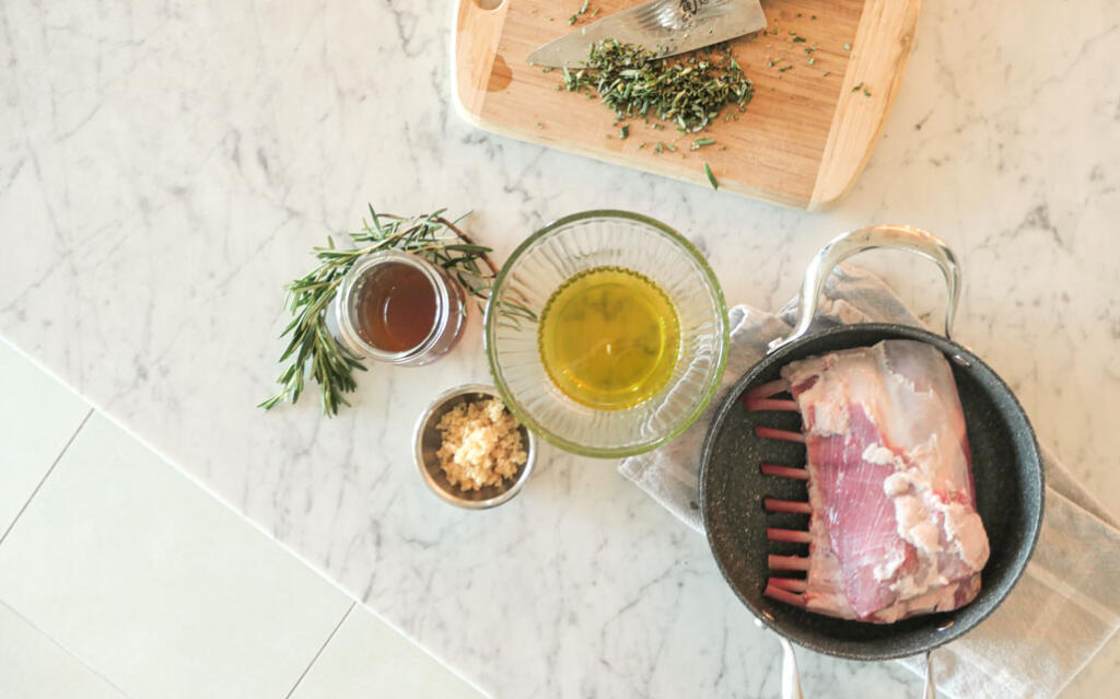 rack of lamb in roasting pan accompanied by ingredients for making roasted rack of lamb on marble counter top