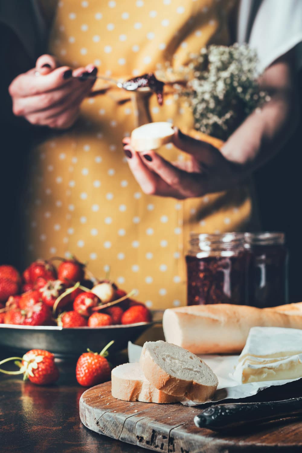 Woman in apron putting strawberry jam onto french bread in front of table with fresh strawberries and cheese and a cutting board