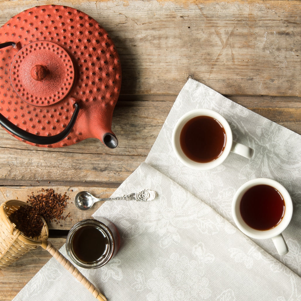 National Hot Tea Month set with red teapot 2 cups filled with hot tea and a tea basket filled with loose leaf tea on wooden table