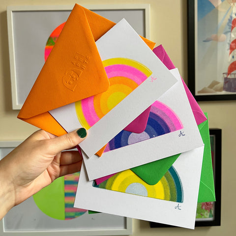 women's hand holding three rainbow cards, a yellow, pink and orange rainbow with an orange envelope, a purple, pink and blue rainbow with a purple envelope and a green and yellow rainbow with a green envelope