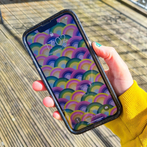 women's hand holding a mobile phone, showing the rainbow wallpaper which is free to download.