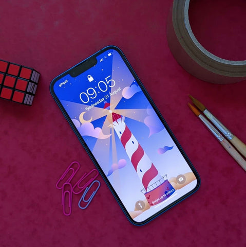 iphone 13 on a red background showing the lighthouse background this months free wallpaper which is red, white, blue and gold. Around the phone there is a roll of tape, paint brushes, paper clips and a tiny rubiks cube