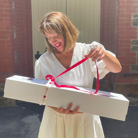 Joy is in the air as she playfully unravels the ribbon, teasing the surprise of the Dozen Ferrari Red Roses & Plush Bunny in a Box, curated with care by Floral Atelier Australia and ready for same day delivery to share the love in Adelaide.