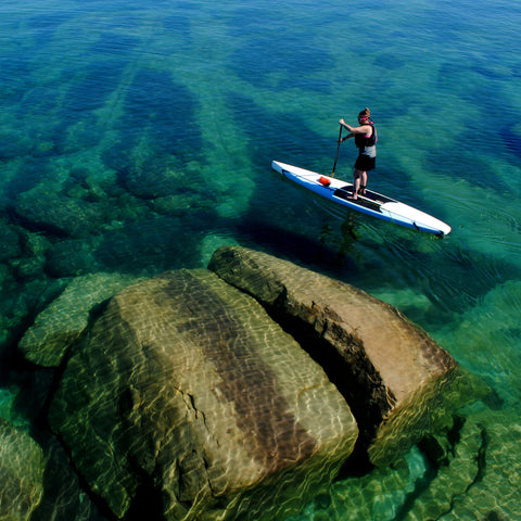 Woman paddles a displacement hull stand up paddleboard above huge rocks on Lake Superior