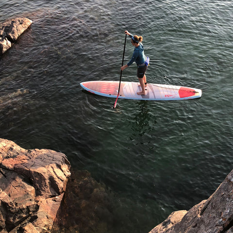 Young woman paddles a hybrid shaped stand up paddleboard near the rocks in Marquette Michigan