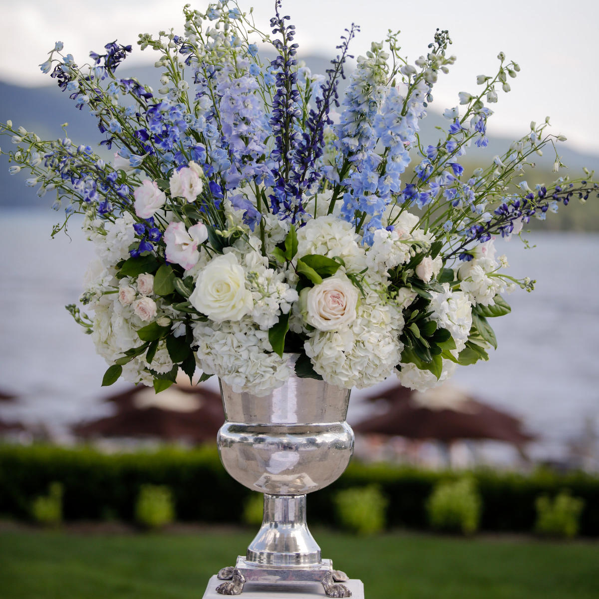 A silver plated floral vesel with stunning wedding flowers, on a white wood pedestal, and a lake view on the background