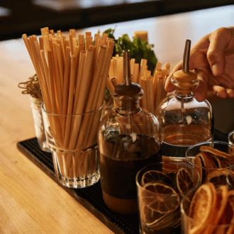 A person's hand reaching for a bottle beside a container of reed drinking straws and citrus garnishes on a bar counter.png__PID:28227b3b-77ee-4c29-8188-20d6e8758dc4