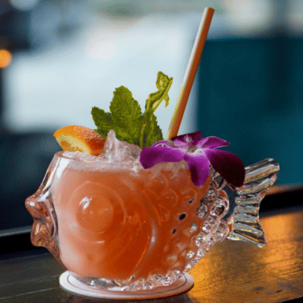 Colorful cocktail in a fish-shaped glass with a Holy City Straw Company straw, garnished with fresh mint, a slice of orange, and a purple orchid, on a bar counter