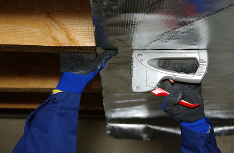 person wearing blue gloves and stapling reflective insulation to a wooden ceiling beam