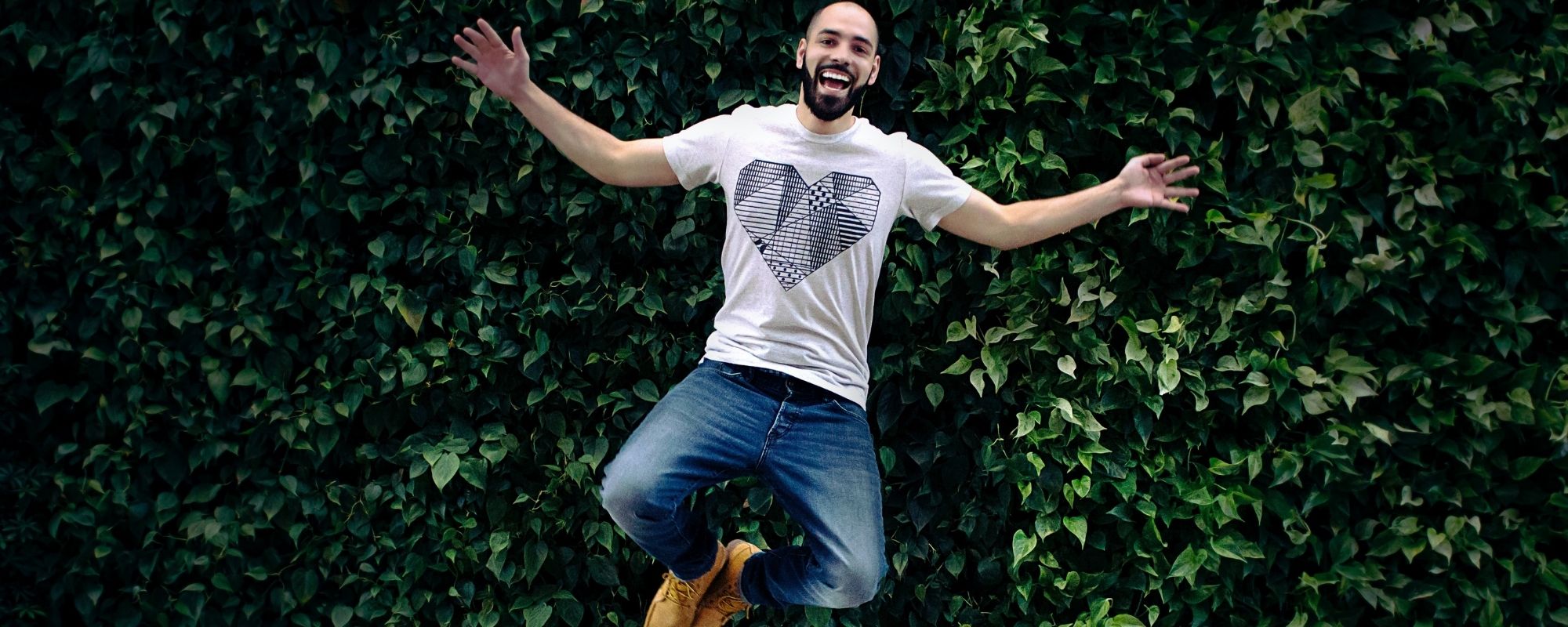 Confident happy Man mid air jumping with arms outstretched against a green headge